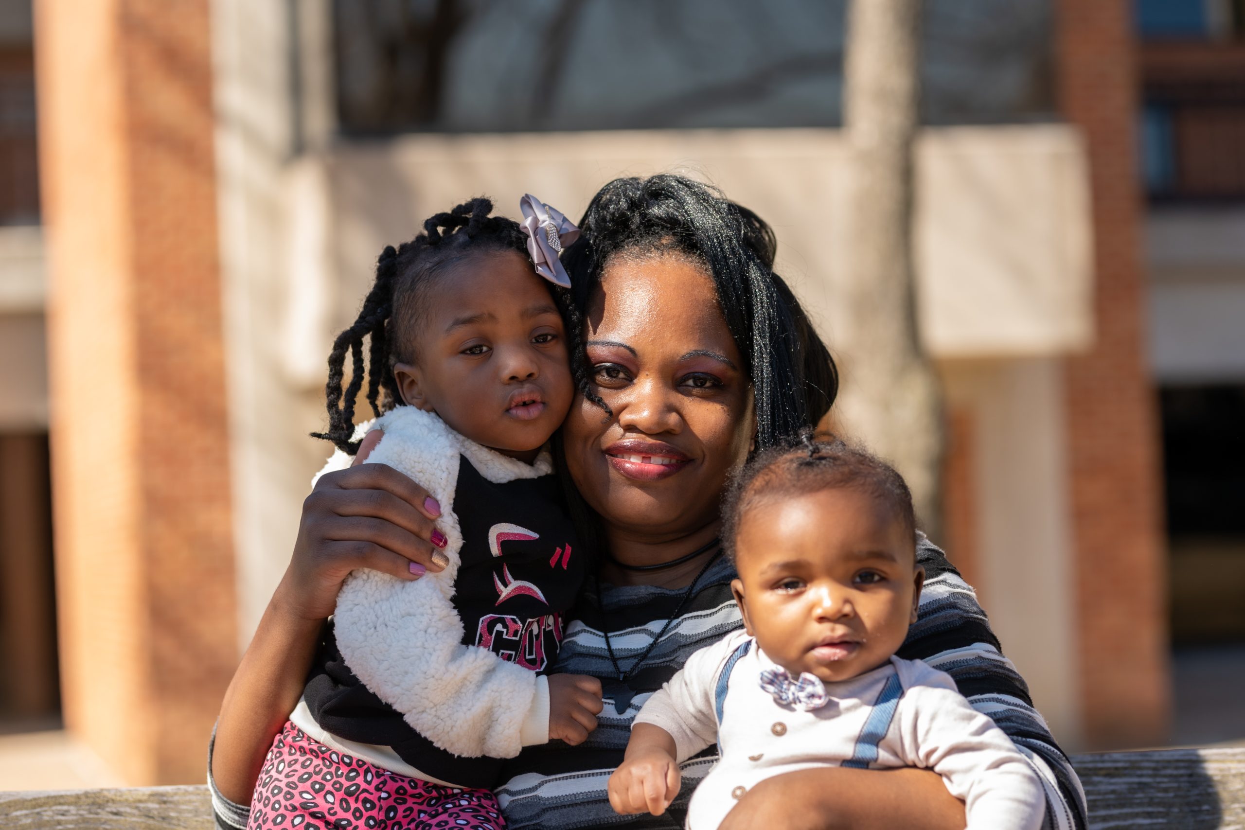 An adult with a dark skin tone is sitting on a bench with their baby with a dark skin tone on their lap and their young child with a dark skin tone standing beside them.