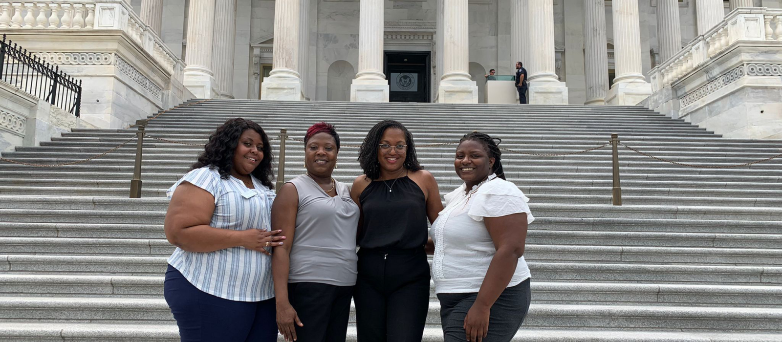 Four adults, all with a dark skin tone, are standing together in front of stairs, posing for a group photo.