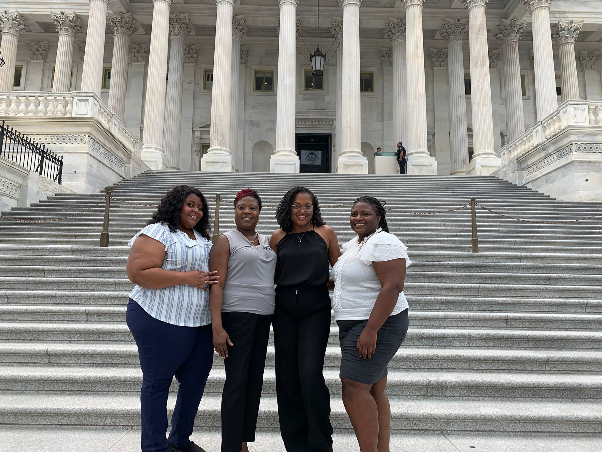 Four adults, all with a dark skin tone, are standing together in front of stairs, posing for a group photo.