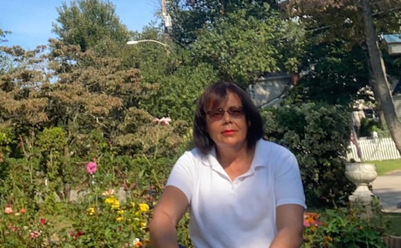 An adult with a medium skin tone is sitting in a garden, posing for a photo with lush greenery behind them.