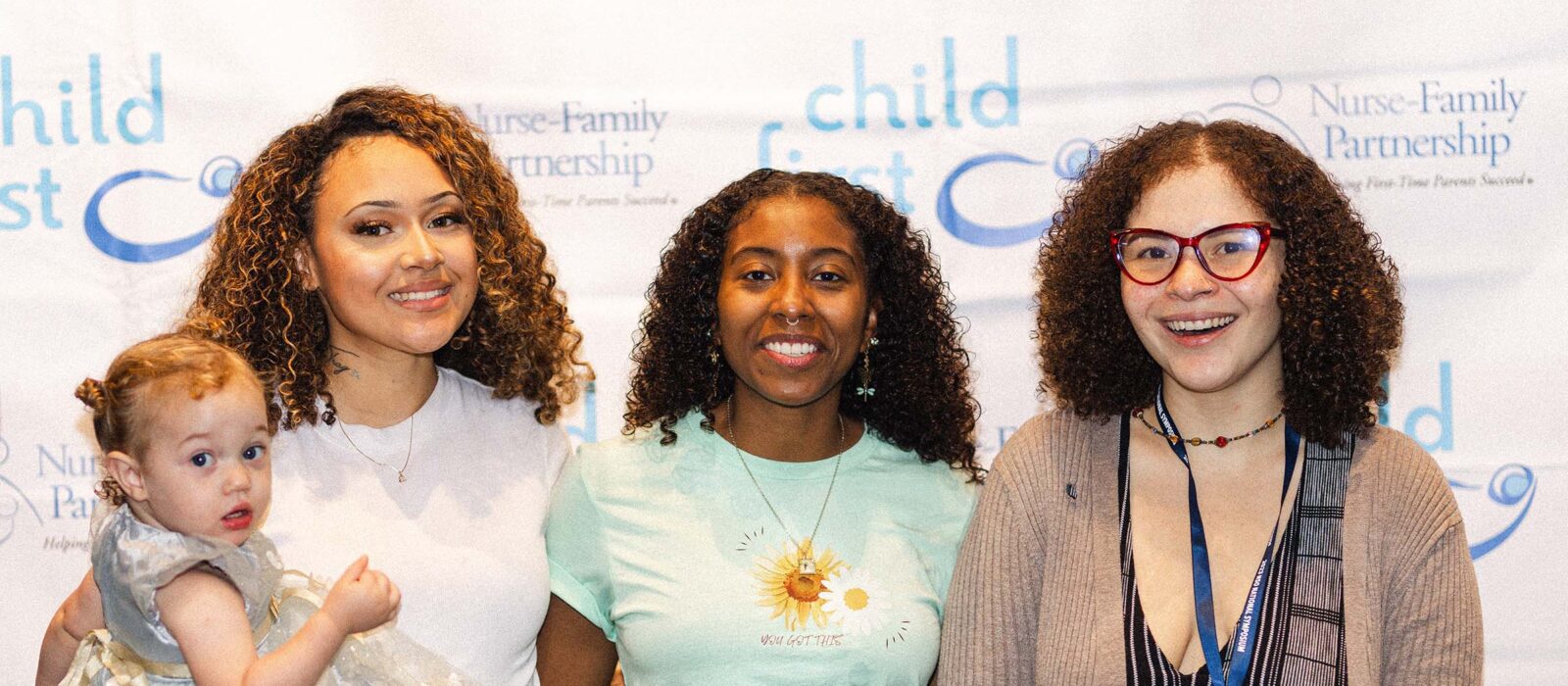 A group of three adults, one with a dark skin tone, one with a light skin tone, and one with a medium skin tone, are posing in front of a Child First and Nurse-Family Partnership backdrop, with one person holding a baby with a light skin tone.