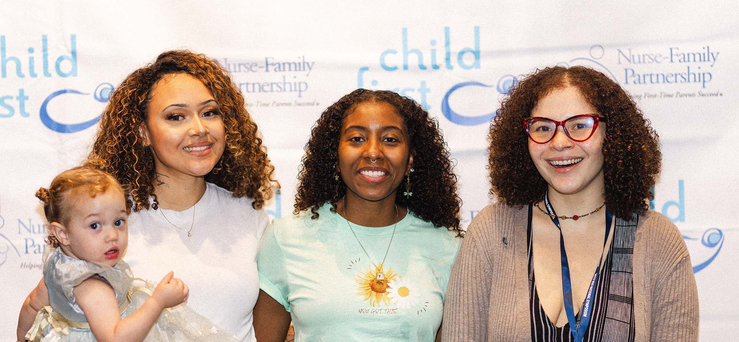 A group of three adults, one with a dark skin tone, one with a light skin tone, and one with a medium skin tone, are posing in front of a Child First and Nurse-Family Partnership backdrop, with one person holding a baby with a light skin tone.