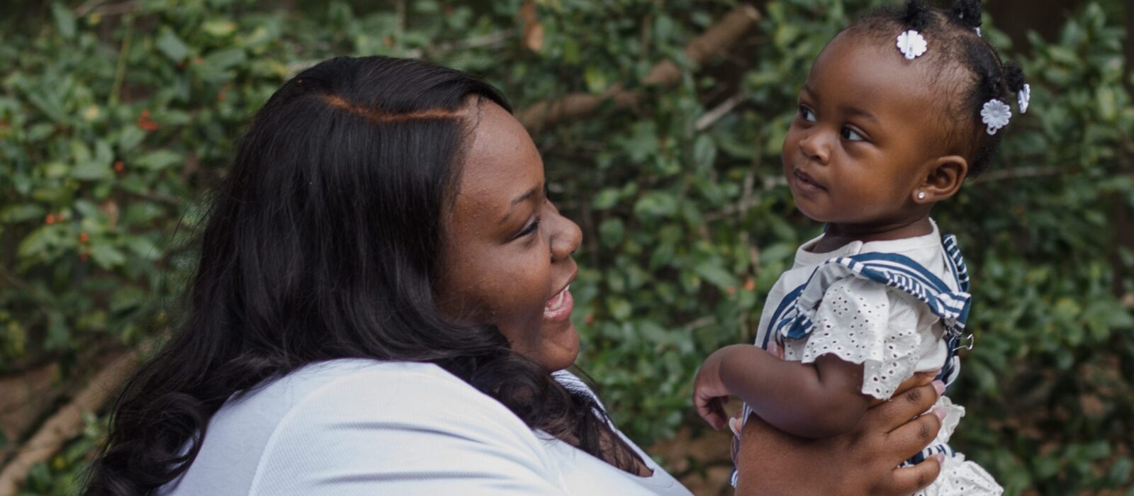 An adult with a dark skin tone is smiling as they lift their baby with a dark skin tone in the air at a park, with greenery in the background.