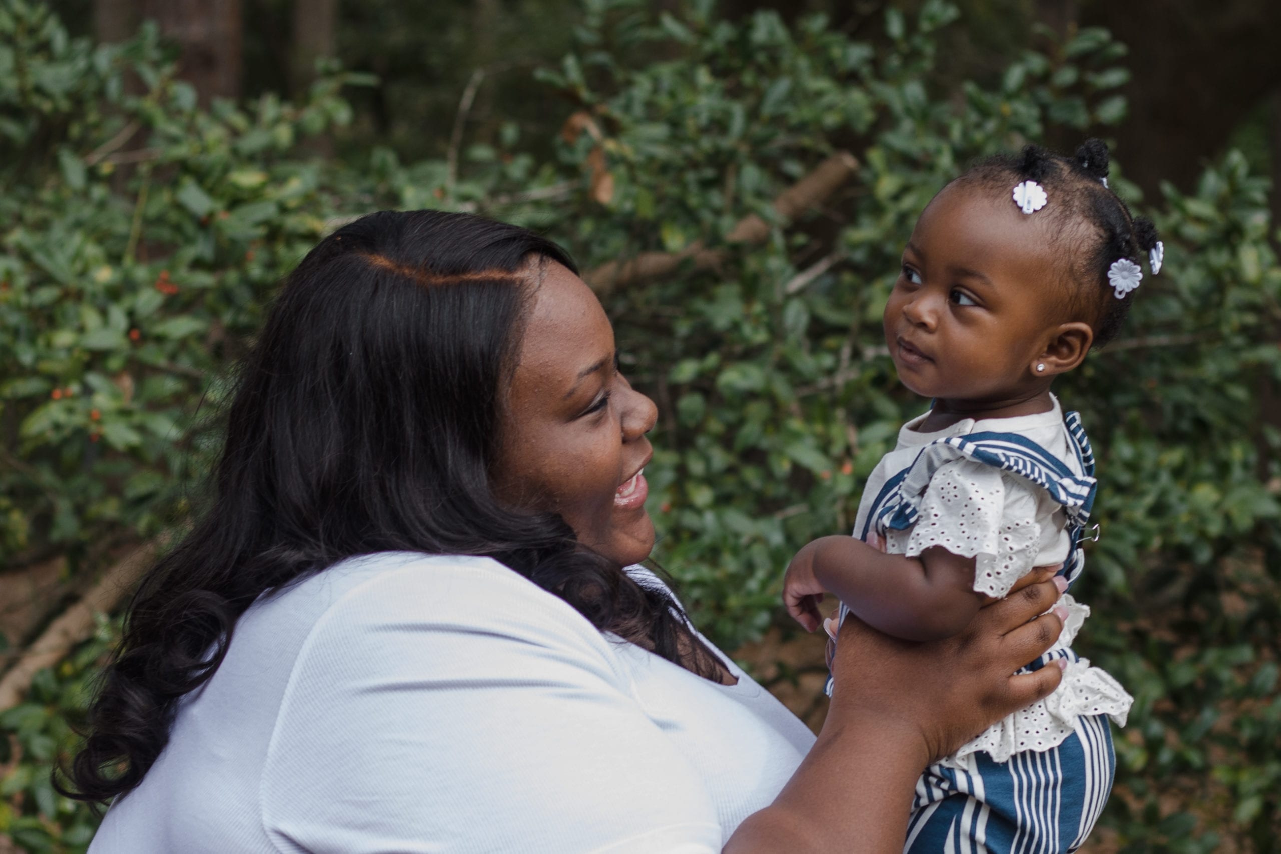 An adult with a dark skin tone is smiling as they lift their baby with a dark skin tone in the air at a park, with greenery in the background.