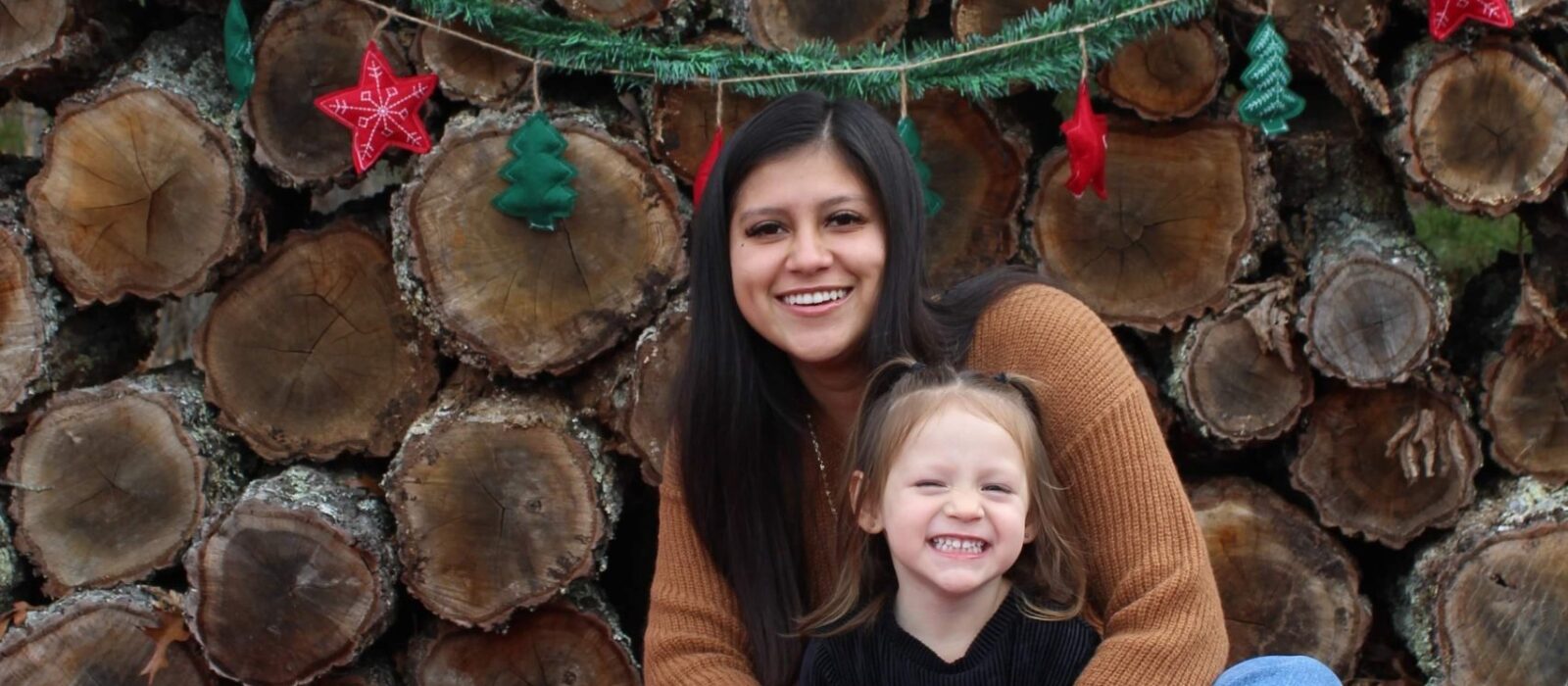 An adult with a medium skin tone is sitting on a flannel blanket with tree logs behind, holding a toddler with a lighter skin tone in their lap.