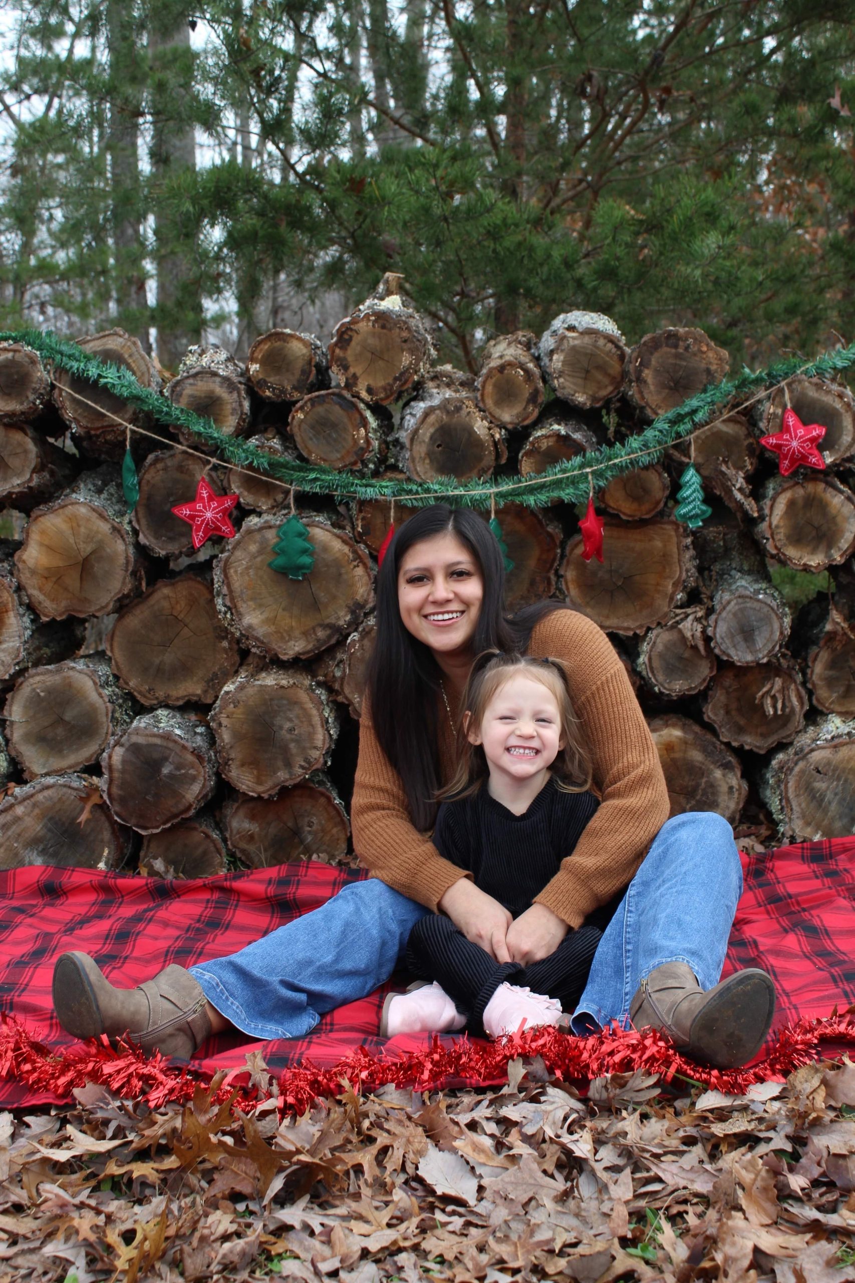 An adult with a medium skin tone is sitting on a flannel blanket with tree logs behind, holding a toddler with a lighter skin tone in their lap.