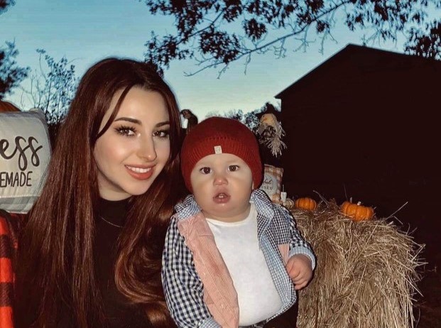 An adult with a light skin tone is sitting in front of a haystack at sunset, holding a baby with light skin on their lap.