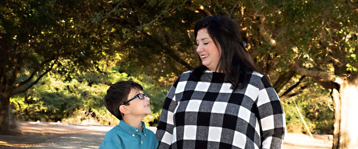 An adult and a child, both with a medium skin tone, are smiling at each other while standing in a tree grove.