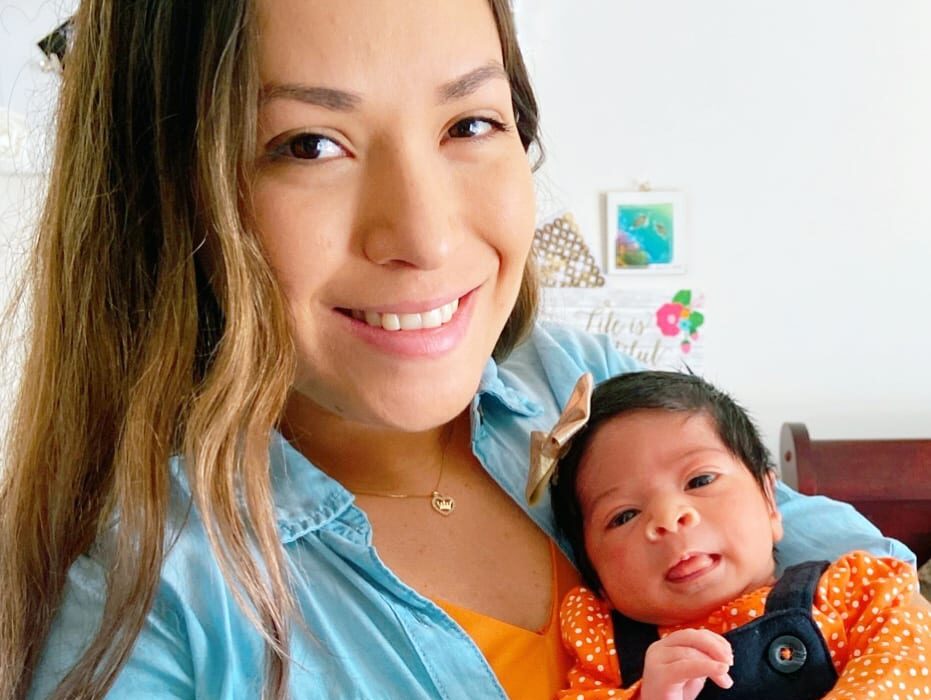 An adult is standing in a bedroom, taking a selfie while holding their infant, both with a medium skin tone.