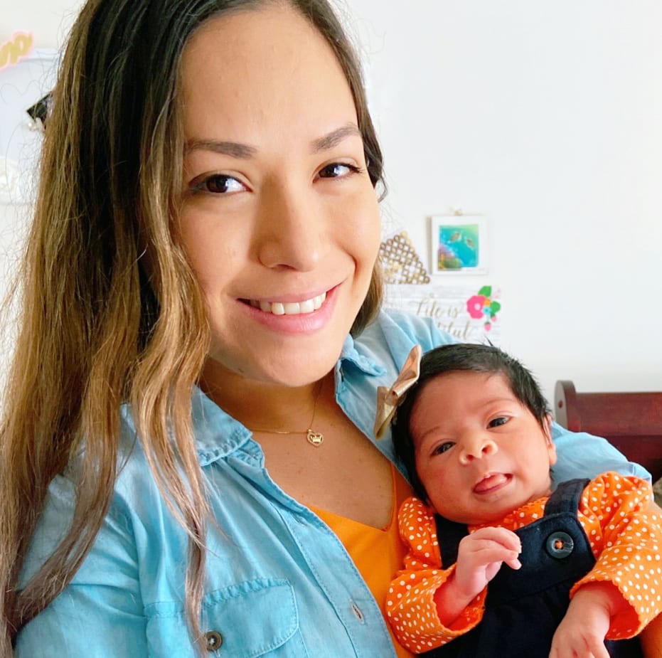 An adult is standing in a bedroom, taking a selfie while holding their infant, both with a medium skin tone.