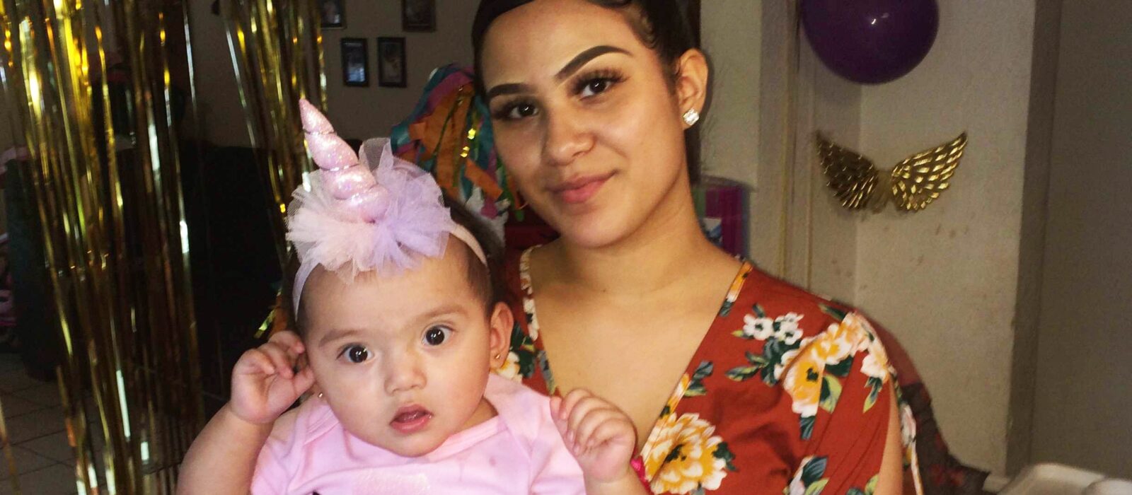 An adult with a medium skin tone is sitting in a decorated kitchen with a baby on their lap, who is wearing a birthday hat.