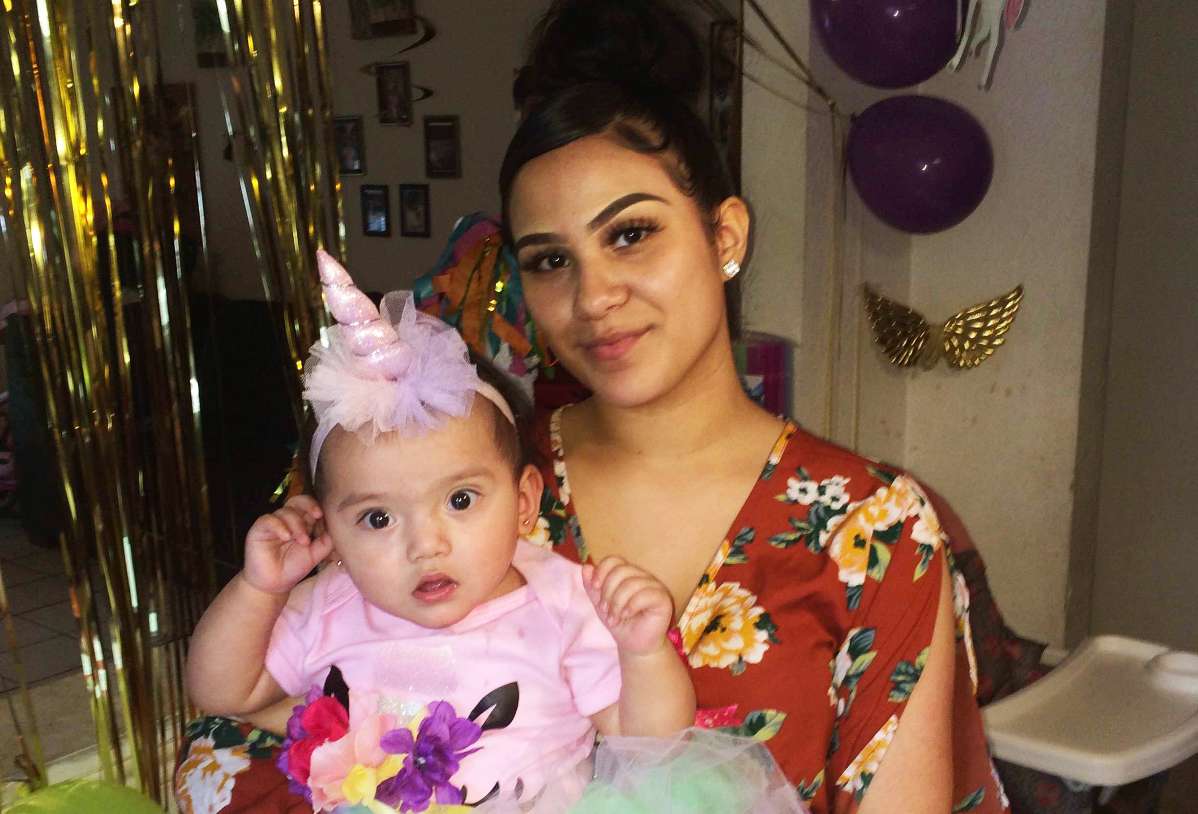An adult with a medium skin tone is sitting in a decorated kitchen with a baby on their lap, who is wearing a birthday hat.