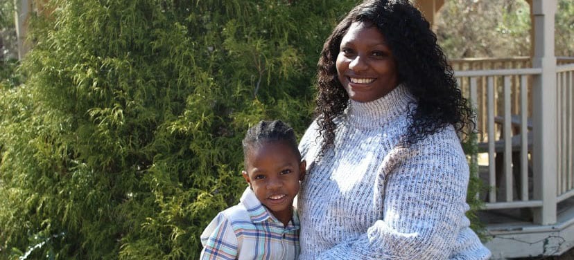 An adult with a dark skin tone is sitting outdoors in front of trees, smiling while posing with a child who has a dark skin tone.