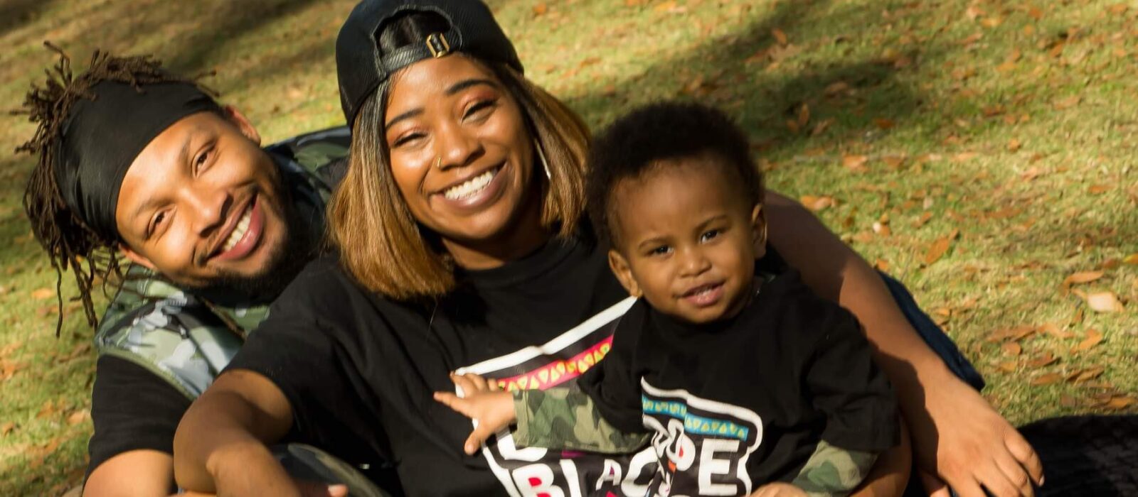 Two adults and a toddler with a dark skin tone sit in the grass at a park together