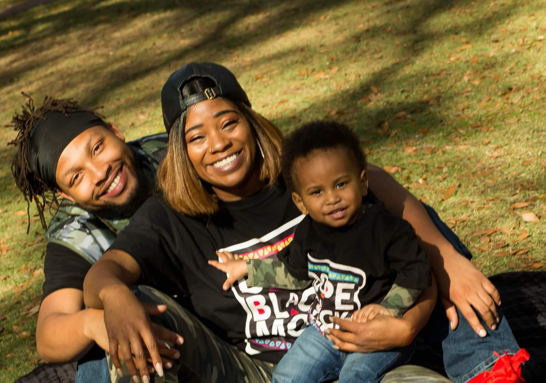 Two adults and a toddler with a dark skin tone sit in the grass at a park together