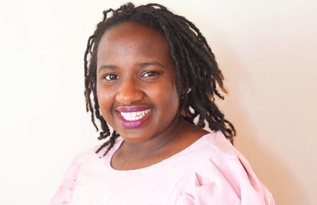 An adult with a dark skin tone is sitting for a portrait in front of a plain, neutral-colored wall.