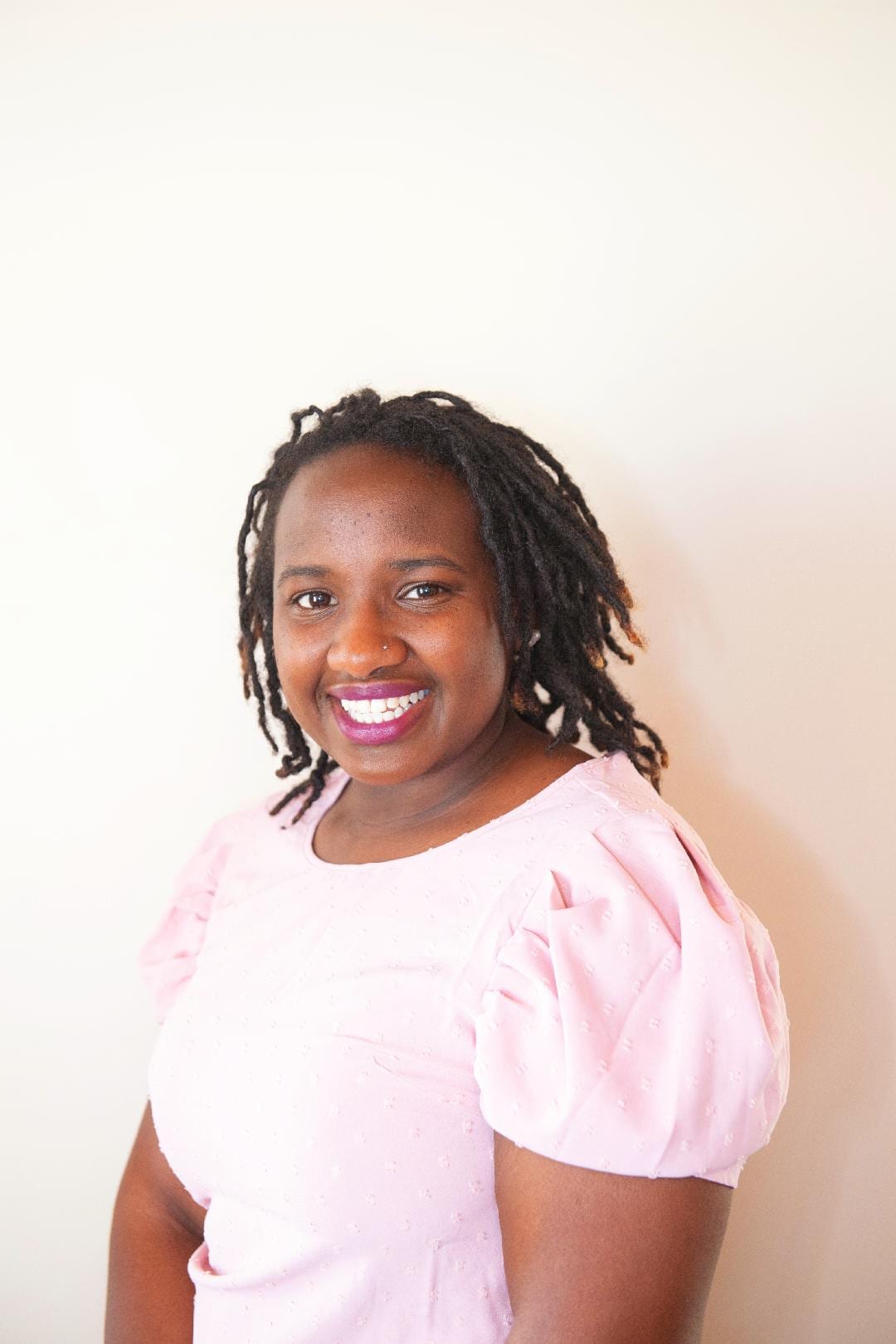 An adult with a dark skin tone is sitting for a portrait in front of a plain, neutral-colored wall.