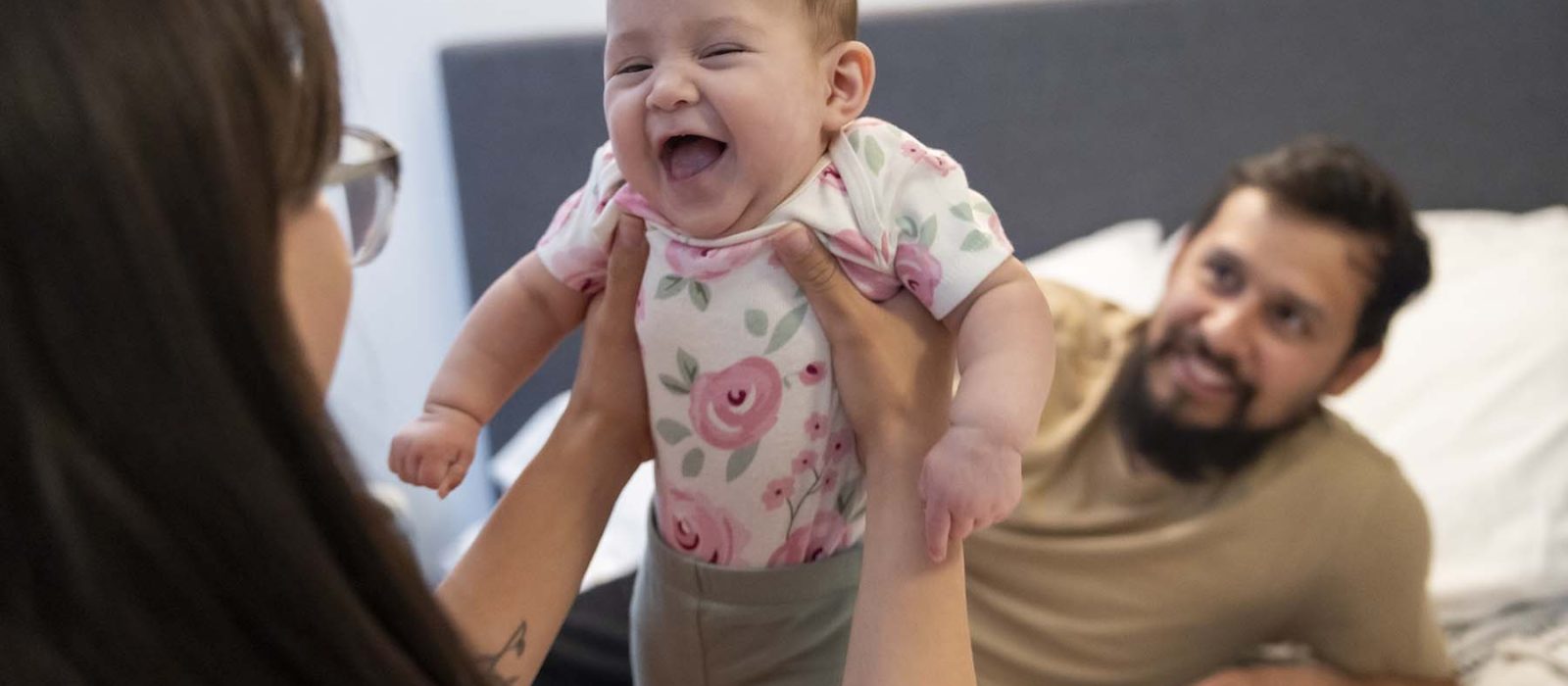 A woman with a medium skin tone lifts a baby playfully into the air while sitting in a bedroom, as a man with a medium skin tone smiles at them from the bed.
