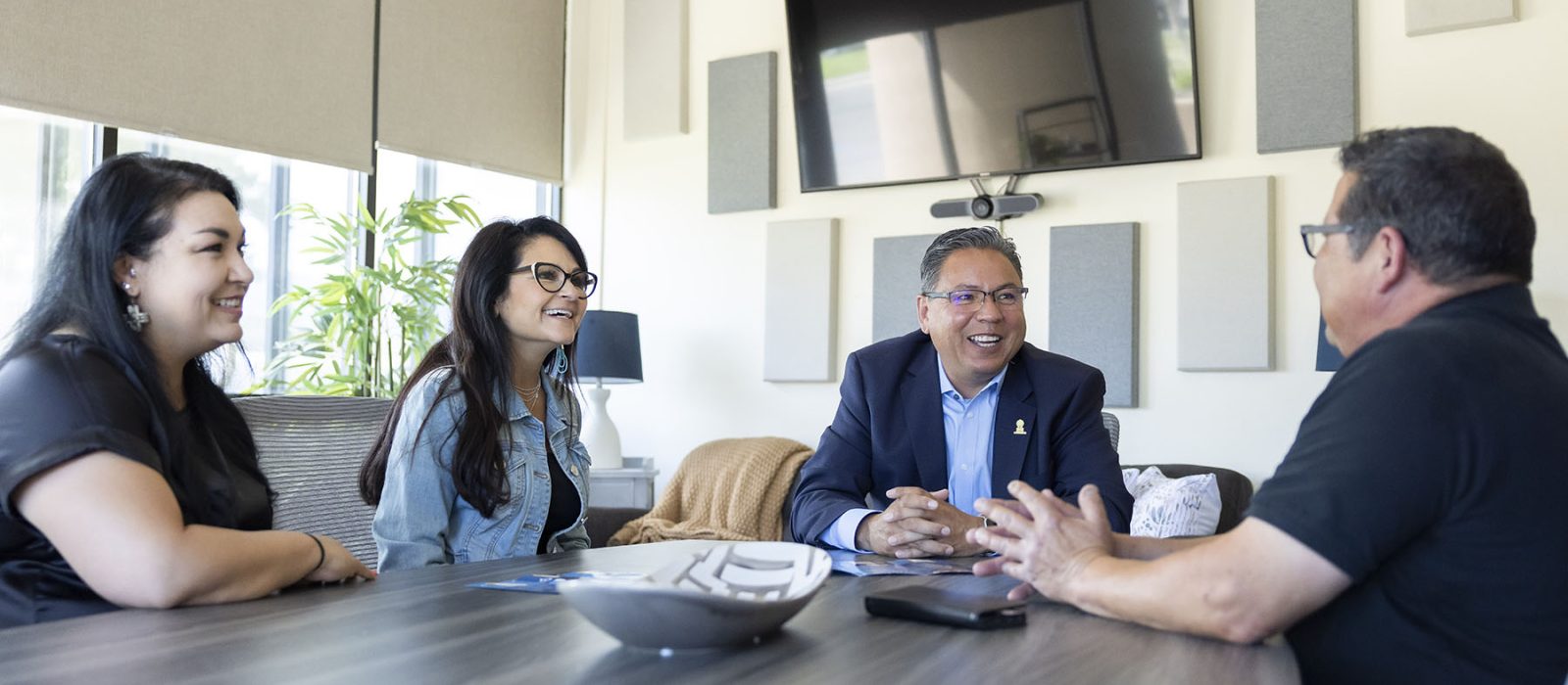 Four adults with medium skin tone sit around a boardroom table, engaged in conversation.