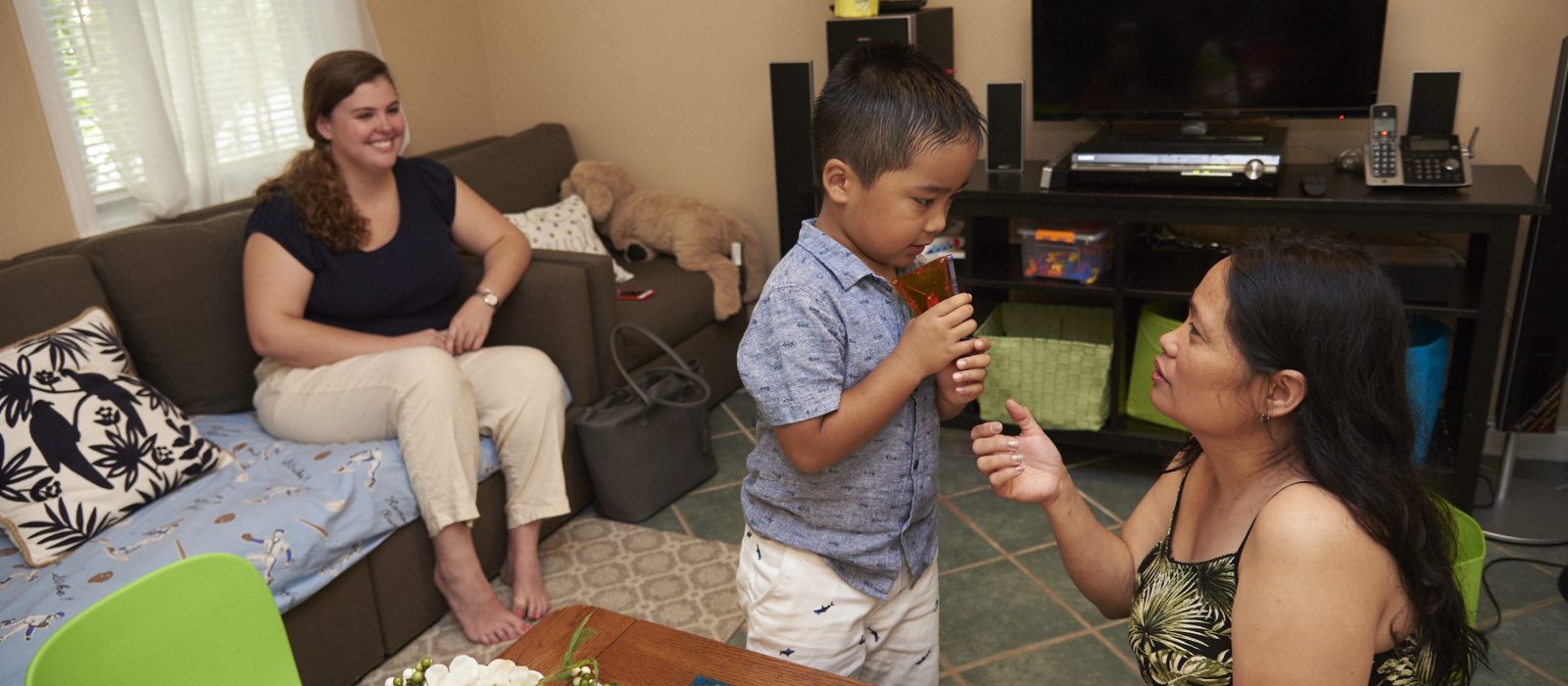 An adult with a light skin tone is sitting on a couch, observing another adult with a medium skin tone sitting on the floor and playing with a toddler with a medium skin tone.