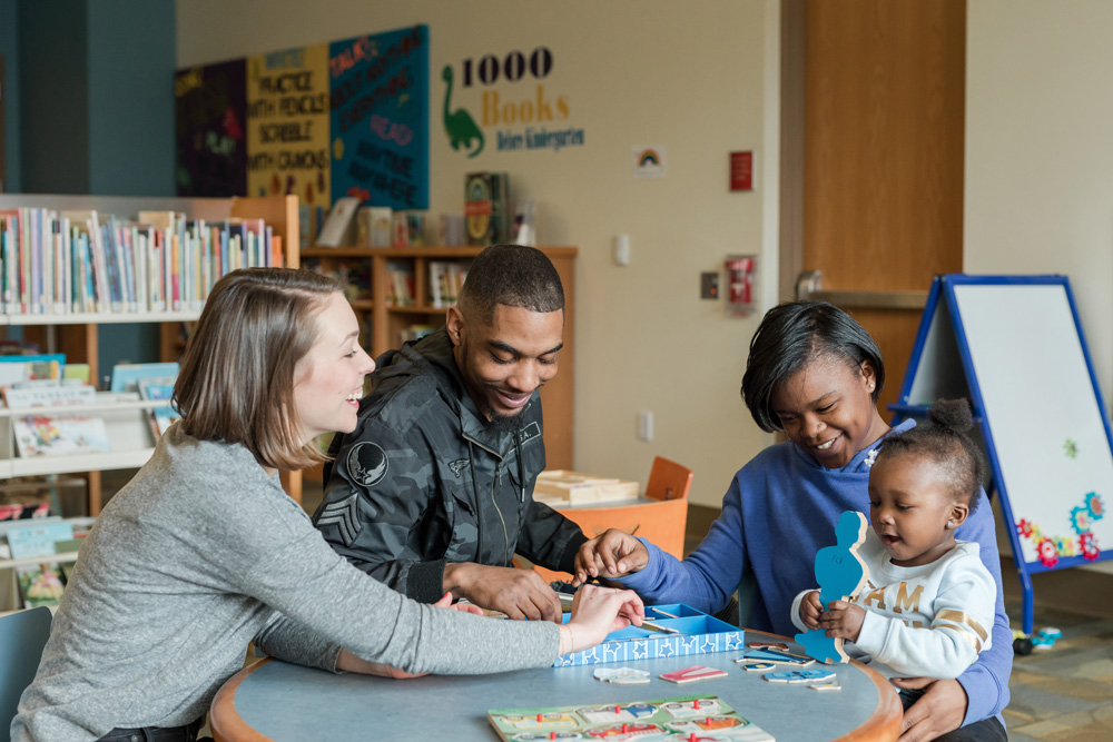 Three adults one with a light skin tone and two with a dark skin tone, sit around a small table, smiling and playing a game with a baby who also has a dark skin tone.