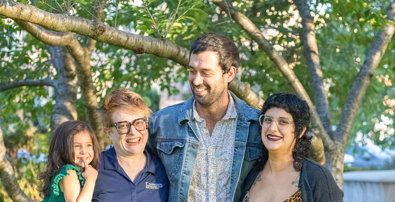 A group of three adults with light skin tones and a child with a light skin tone are posing for a photo in front of some trees, smiling together.