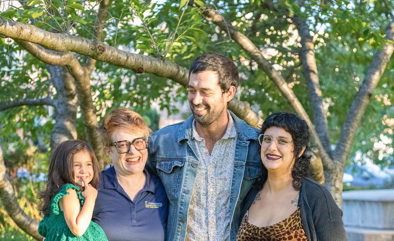 A group of three adults with light skin tones and a child with a light skin tone are posing for a photo in front of some trees, smiling together.