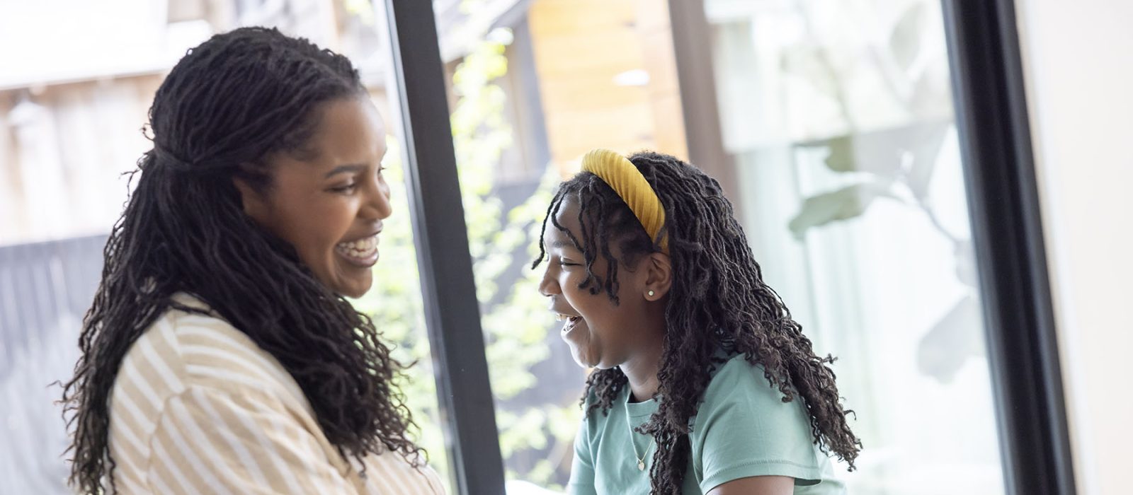 A woman and a young girl, both with dark skin tone, sit together in front of a set of windows, laughing and enjoying each other’s company.