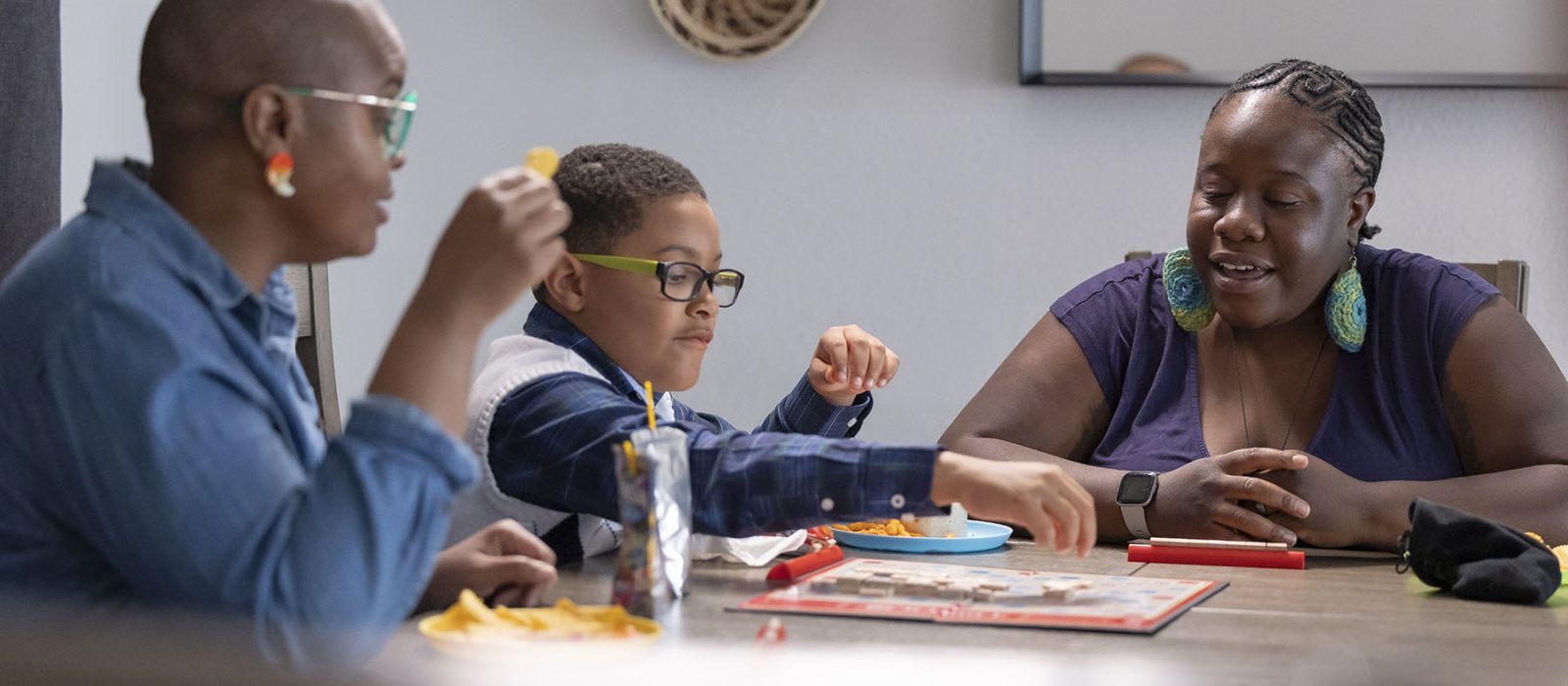 Two women and a young boy with a dark skin tone, sit at a kitchen table, enjoying snacks and playing a game together.