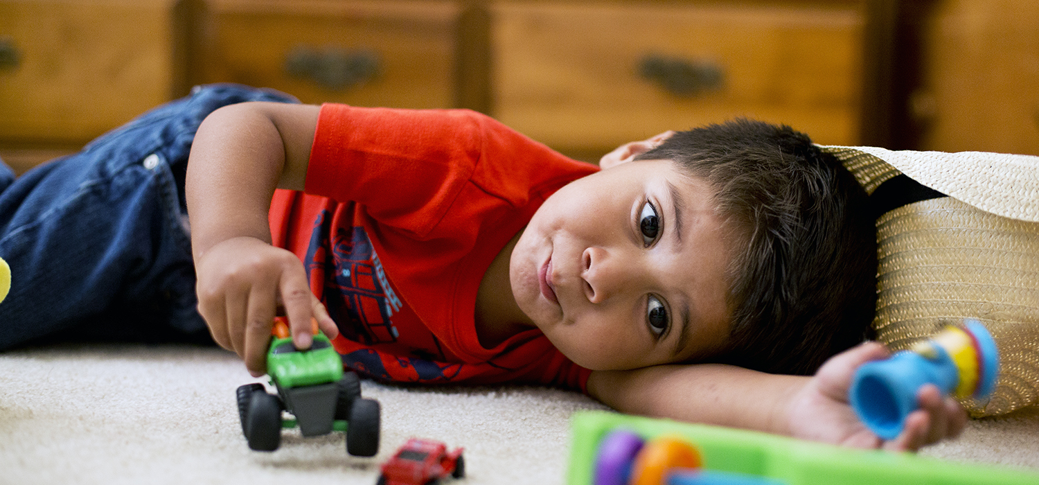 A baby with a medium skin tone is lying on the floor in a living room, playing with a toy truck, surrounded by other toys.