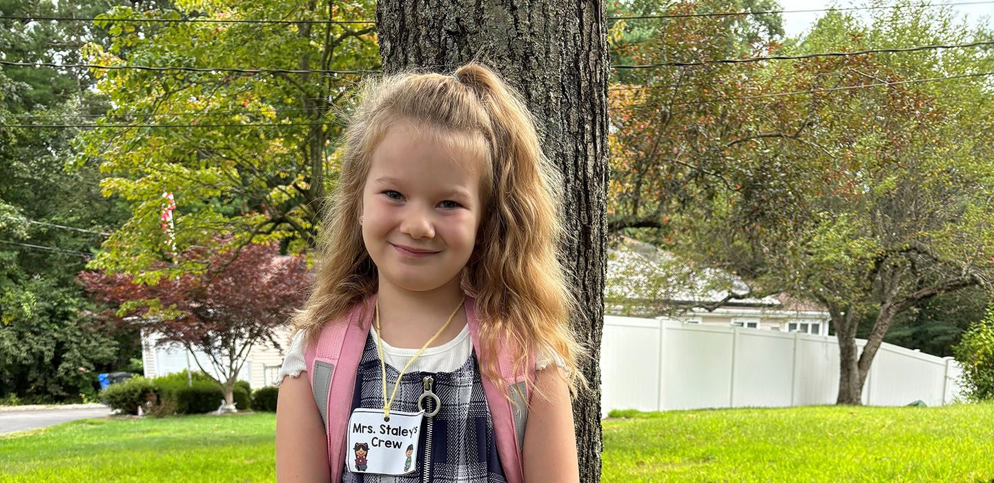 A child with a light skin tone is standing in front of a tree, wearing a backpack with their hair styled half-up, half-down.