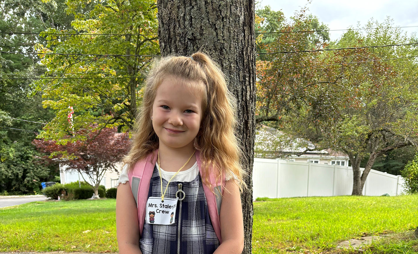 A child with a light skin tone is standing in front of a tree, wearing a backpack with their hair styled half-up, half-down.