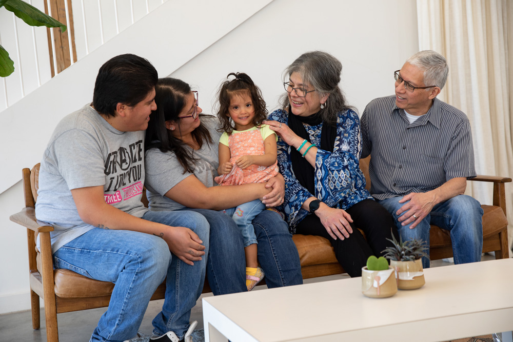 Four adults with medium skin tones sit on a couch in a living room, smiling and watching a toddler with a medium skin tone seated in the middle.
