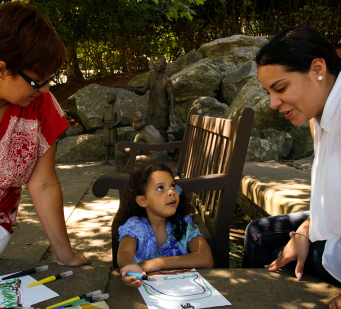Two adults with a medium skin tone are sitting together at a table with a child who has a medium skin tone, looking at a drawing.