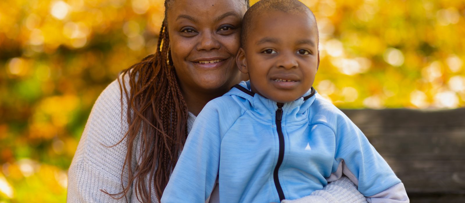 An adult with a dark skin tone is sitting on a park bench, smiling while posing with their child who has a dark skin tone.