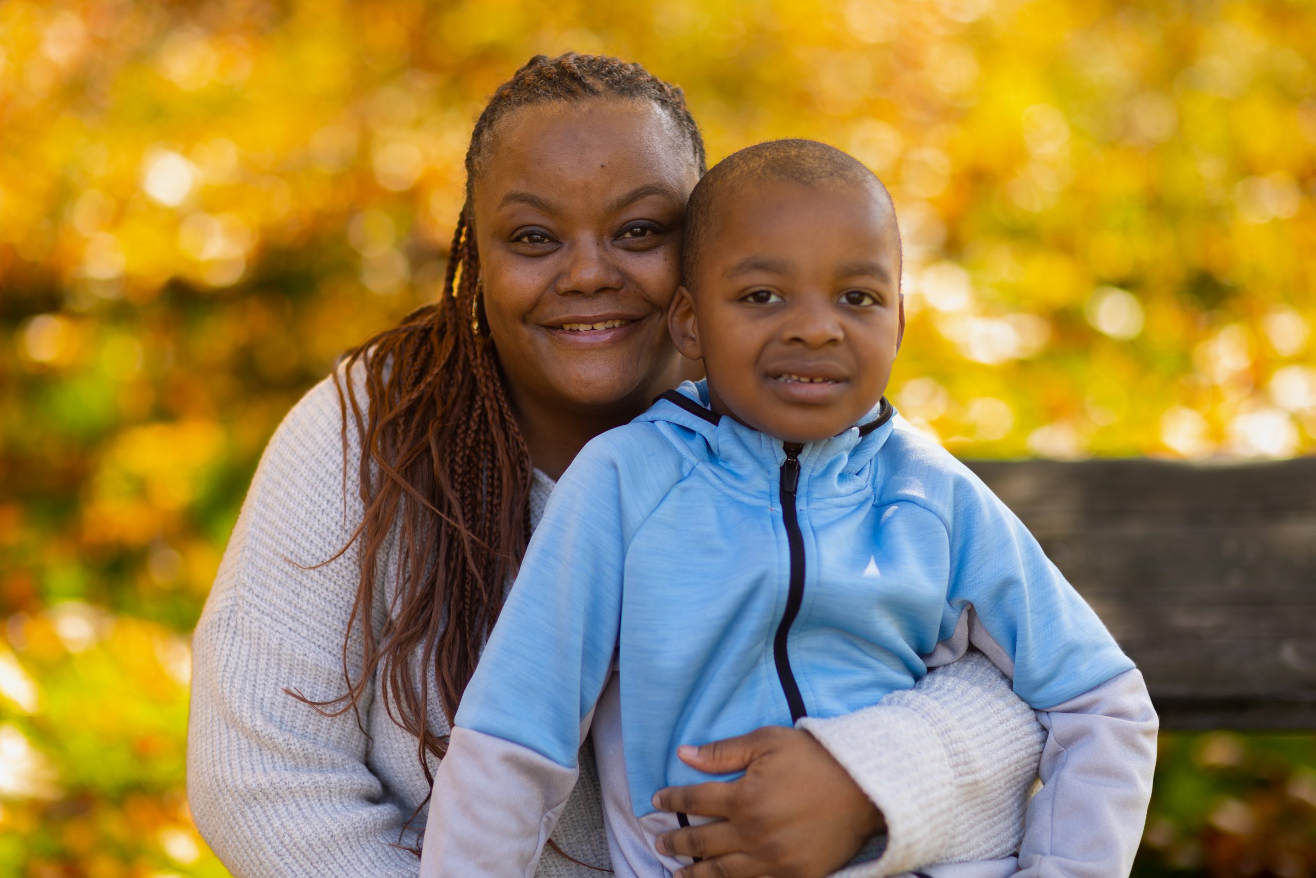 An adult with a dark skin tone is sitting on a park bench, smiling while posing with their child who has a dark skin tone.