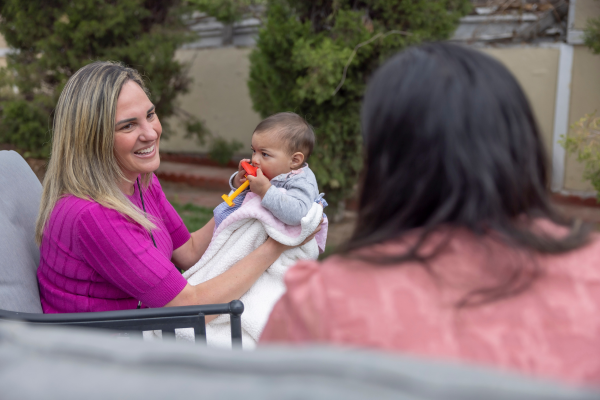 A female Nurse-Family Partnership staff member with a light skin tone holds a baby in her lap while speaking with the baby’s mother.. The mother and baby have a medium skin tone.